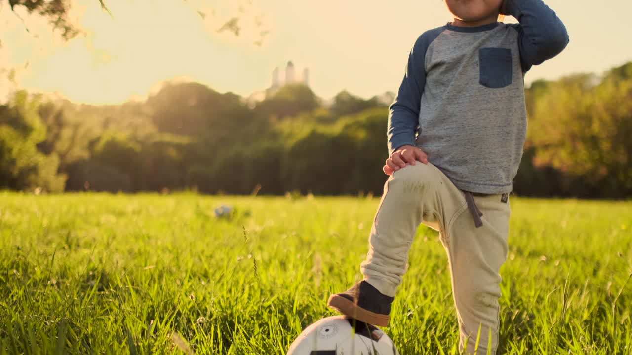un niño de pie en la hierba con una pelota de fútbol al atardecer.