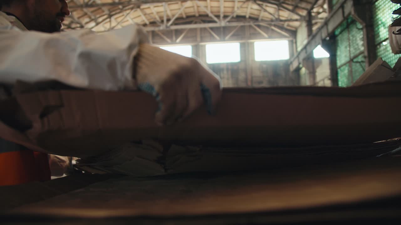 um cara moreno com barba em um uniforme branco e um colete laranja empilha folhas de papelão junto com seus colegas em pilhas especiais em uma fábrica de reciclagem de resíduos