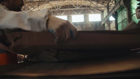 a brunette guy with a beard in a white uniform and an orange vest stacks sheets of cardboard together with his colleagues in special piles at a waste recycling plant