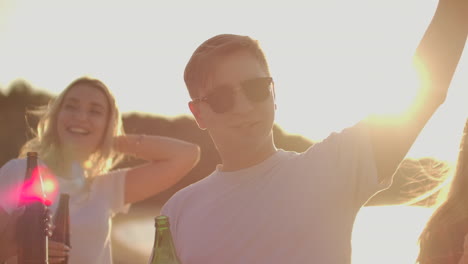 Un-Joven-Con-Camiseta-Blanca-Y-Gafas-De-Sol-Negras-De-Moda-Mueve-Las-Manos-Y-Baila-Con-Sus-Mejores-Amigos-En-La-Fiesta-Al-Aire-Libre-Con-Cerveza.-Disfruta-De-Esta-Tarde-De-Verano-Al-Atardecer.