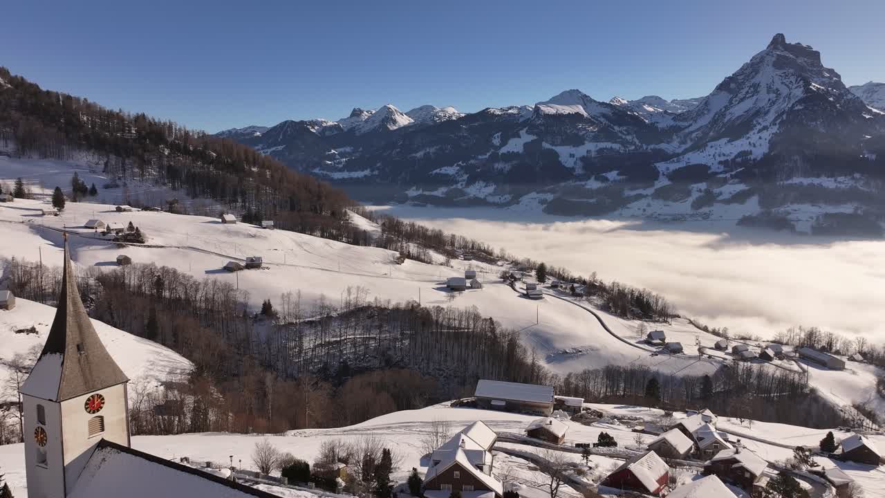 Aerial view of Gallus Church in Amden, Switzerland, surrounded by a snow-covered landscape. The church stands tall against the backdrop of towering mountains and misty valleys.