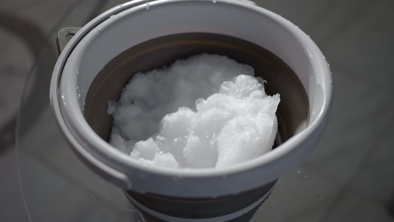 Collapsible bucket filled with frozen water and ice crystals placed on round glass table in indoor space close up shot showing texture and reflection in soft diffused light with gentle shadows
