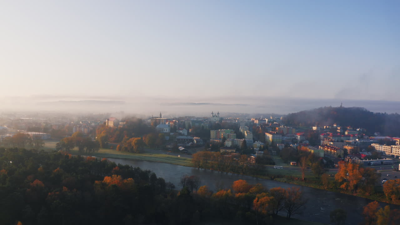 Drone shot of small town covered in early morning fog, buildings and trees bathed in soft, warm sunlight. Mist creates a dreamy effect over the landscape, blue sky above adds a sense of calmness.
