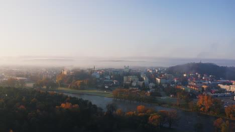 drone shot of small town covered in early morning fog, buildings and trees bathed in soft, warm sunlight. mist creates a dreamy effect over the landscape, blue sky above adds a sense of calmness.