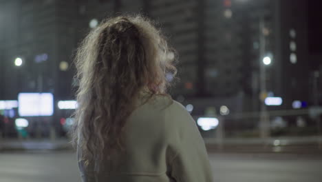 back view of lady standing outdoors late at night wearing white coat under streetlights with soft bokeh glow from city buildings and illuminated windows in urban setting