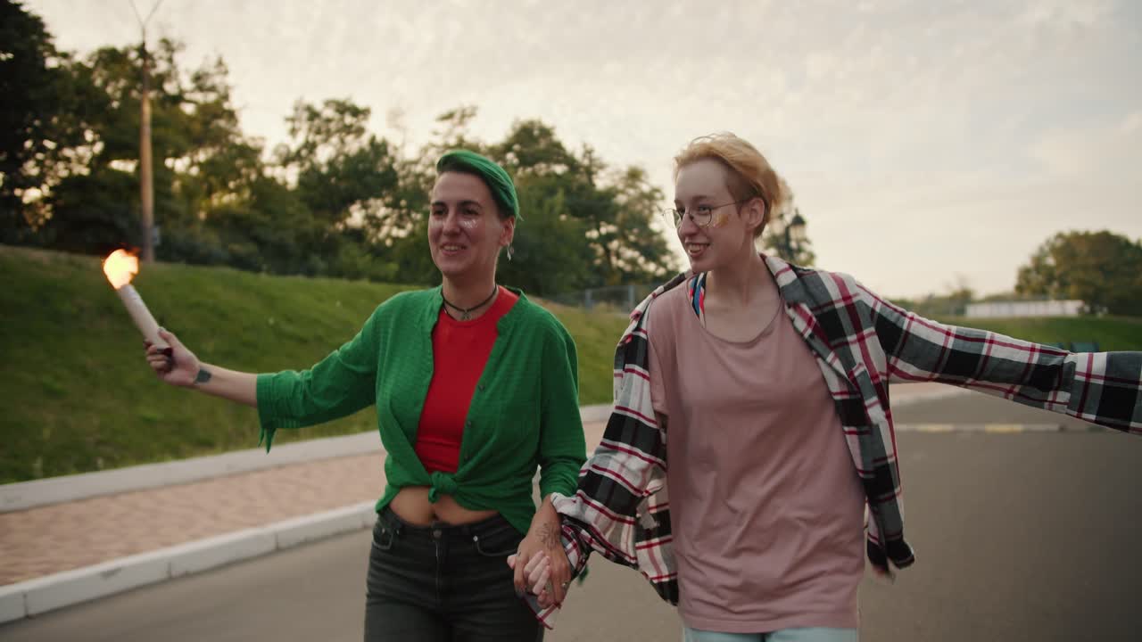 Two lesbian girls with short bright haircuts in plaid shirts run and hold fireworks in their hands during their date in the park in the summer
