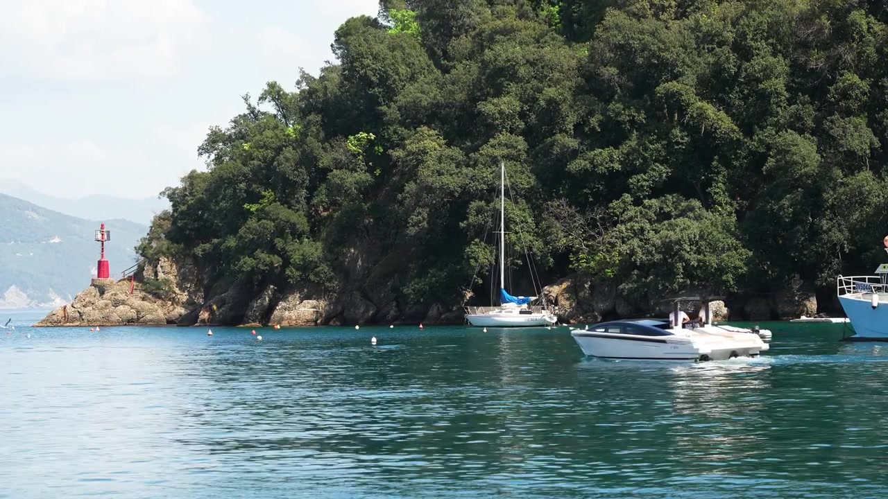 Ampia inquadratura diurna di barche che scivolano sulle acque blu scintillanti vicino a Portofino, Italia, con fitte colline verdi, costa rocciosa e un faro rosso in lontananza. Nessuna persona visibile