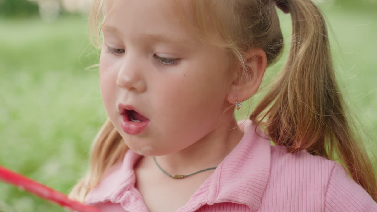 Caucasian girl blowing bubbles in meadow closeup portrait shows delicate pigtails, pink shirt, soap wand, tiny soapy bubbles forming on lips, soft sunlight, gentle breeze, serene summer atmosphere