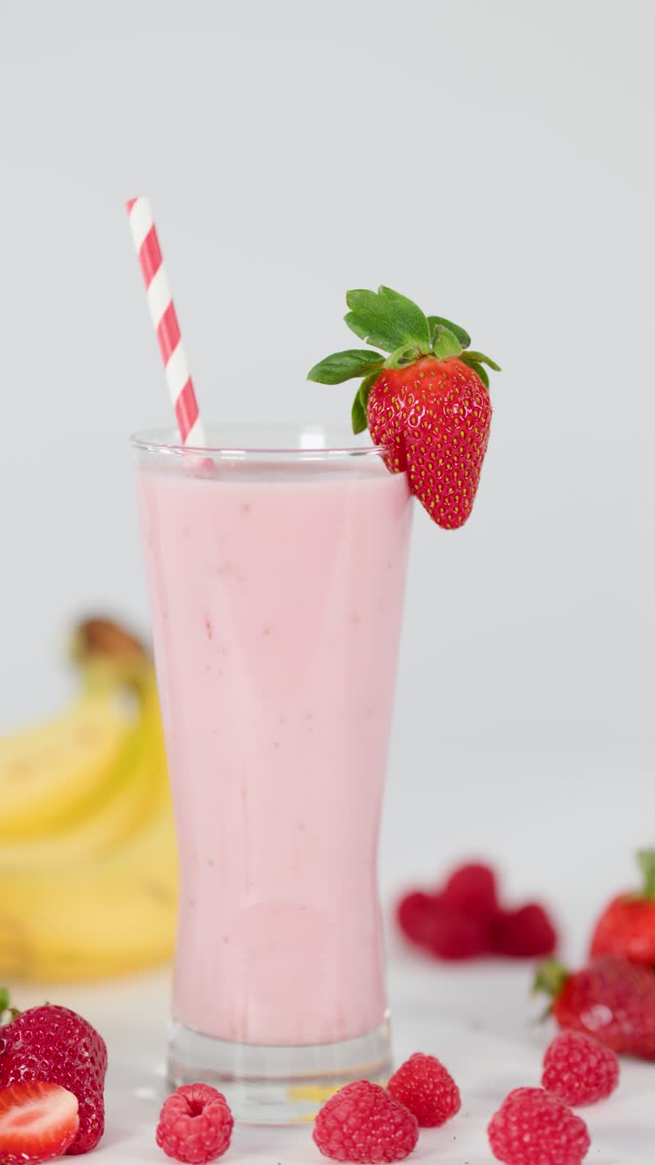 Smoothie rosa cremoso in un bicchiere alto, guarnito con una fragola, circondato da banane, ananas e lamponi.