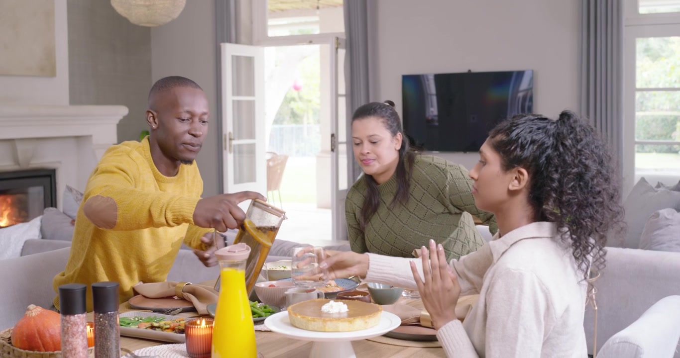 Família afro-americana aplaudindo enquanto mulher entra com tigela, homem serve jarra na mesa de casa, compartilhando.