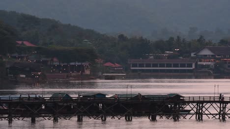 Mon-Bridge-and-a-longboat-followed-speeding-behind-the-structure-while-people-are-walking-on-the-bridge,-silhouetting-as-it-was-getting-dark,-in-slow-motion