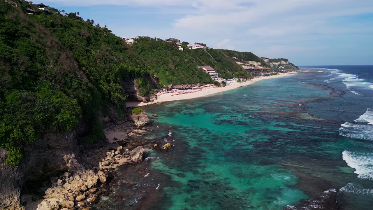 Aerial footage of Melasti Beach Bali showcasing turquoise coastal waters, coral formations, and towering cliffs along a pristine tropical shoreline bathed in warm sunlight and calm ocean breeze