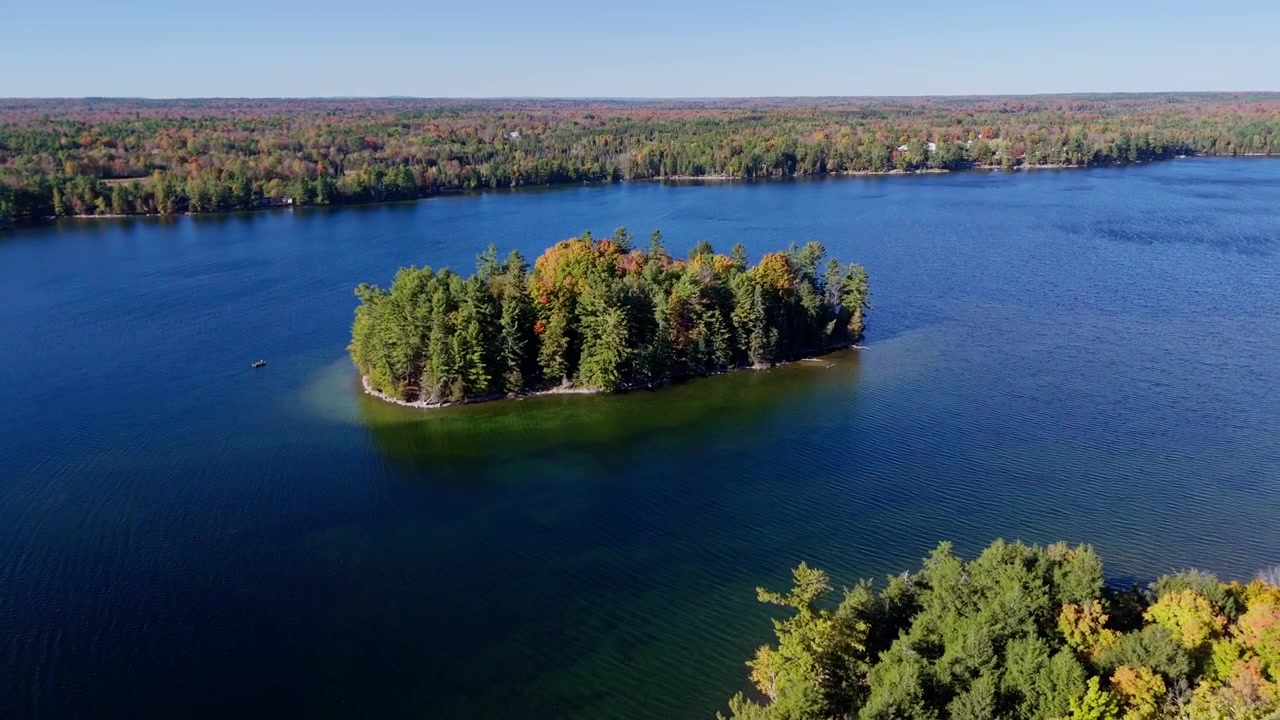 Imagem cinematográfica de drone voando para trás e para cima para revelar uma pequena ilha com um barco ancorado nas proximidades. Folhagem vibrante de outono e natureza selvagem intocada do lago em Ontário.