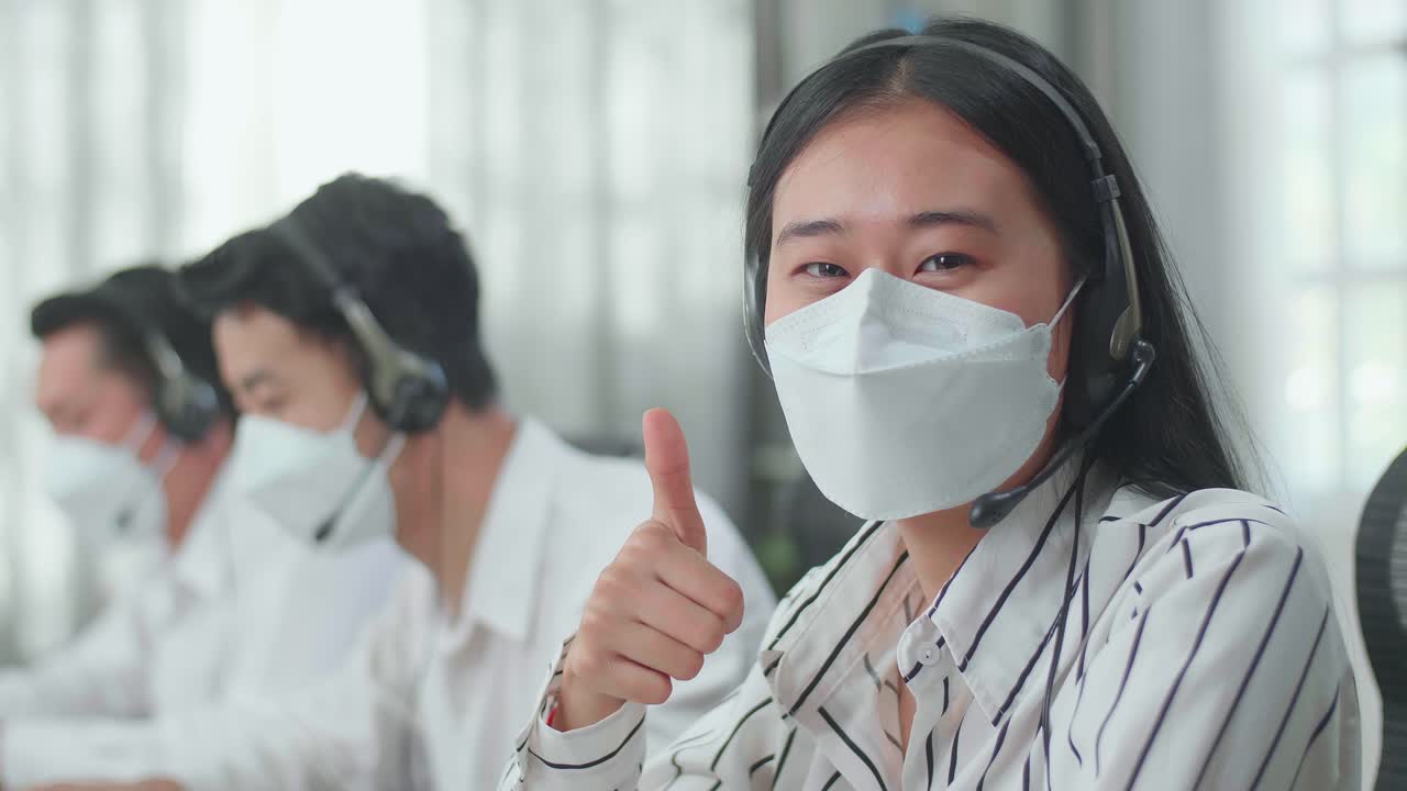 Close Up Of A Woman Of Three Asian Call Center Agents Wearing Headset And Mask Looking At Camera And Thumbs Up While Two Of Her Colleagues Are Speaking With Customer At The Office