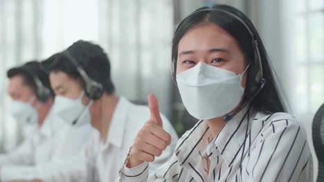 close up of a woman of three asian call center agents wearing headset and mask looking at camera and thumbs up while two of her colleagues are speaking with customer at the office