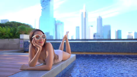 Asian-girl-with-big-sunglasses-sunbathing-lying-on-pool-edge-and-skyscrapers-in-background