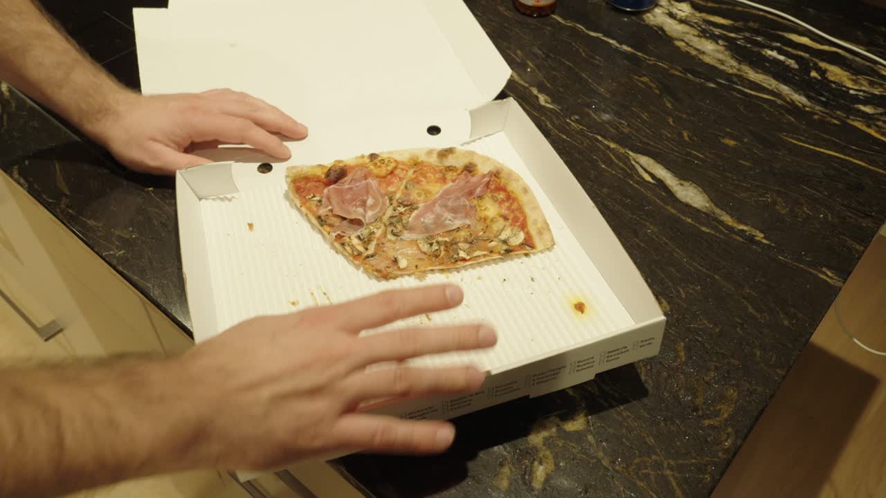 A hand opens a pizza box revealing leftover slices, showing food waste, snack time or late night eating