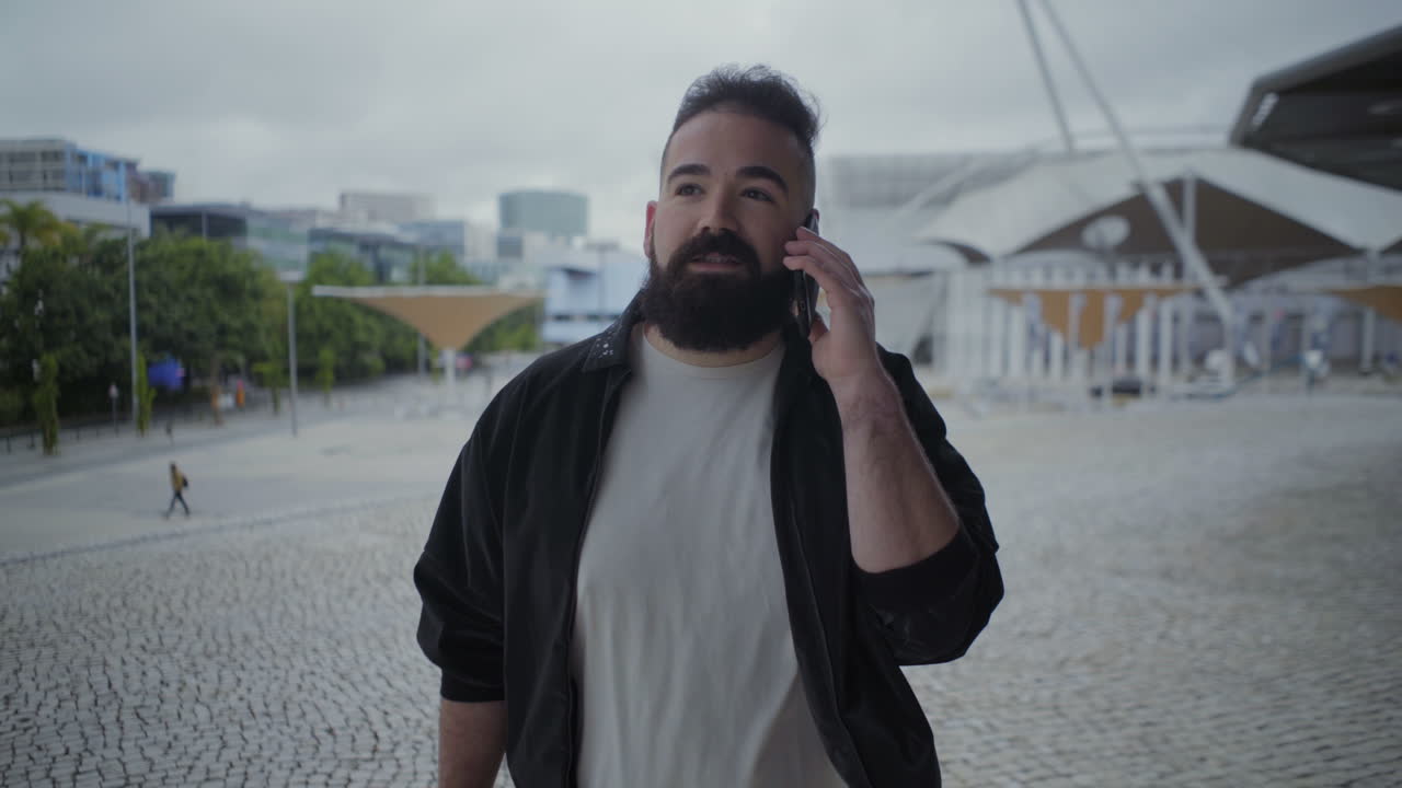 веселый бородатый парень, идущий по площади со смартфоном.