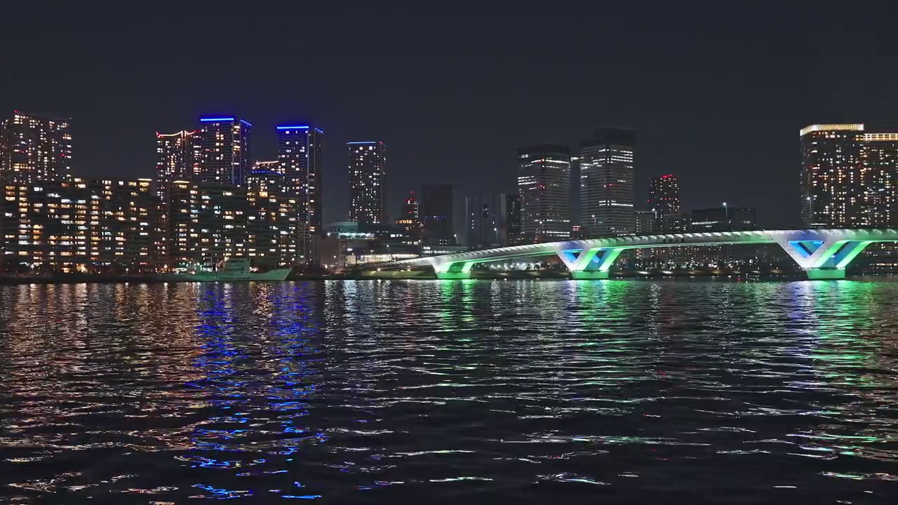 Wide static shot of Tokyo Bay at night, dense glittering high-rise towers reflecting colorful lights on rippling dark water, Harumi Bridge green and blue illumination visible right of frame.