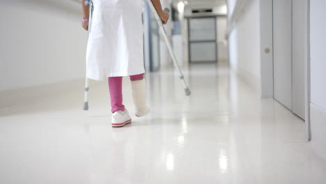 lowsection of african american girl in hospital gown walking using crutches in corridor, slow motion