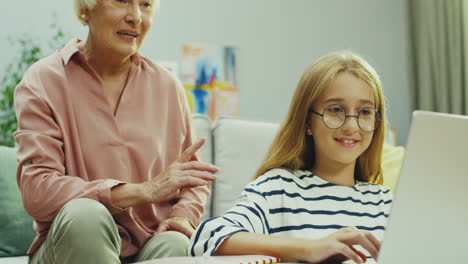 Porträtaufnahme-Des-Hübschen-Teenager-Mädchens-Mit-Brille,-Das-Etwas-Auf-Dem-Laptop-Computer-Tut,-Und-Ihrer-Großmutter,-Die-Redet-Und-Ihr-Sagt,-Was-Sie-Tun-Soll,-Während-Sie-Auf-Den-Bildschirm-Schaut