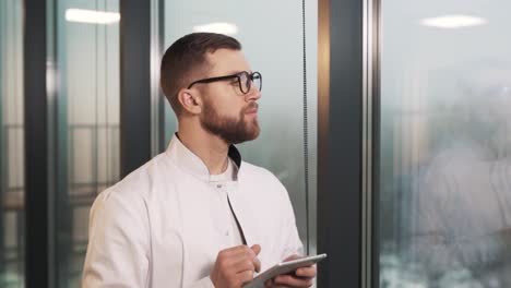 Portrait-of-a-male-doctor-in-a-white-coat-with-a-tablet-standing-near-panoramic-windows-in-a-modern-new-clinic