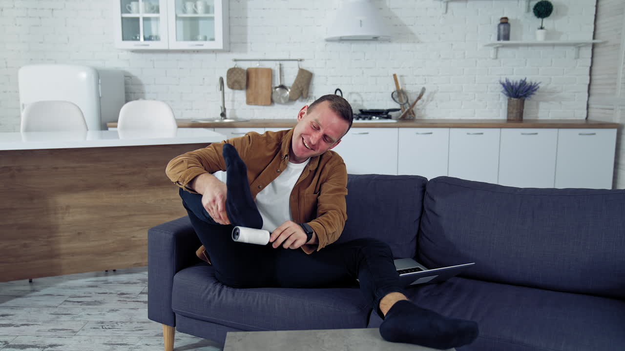 Young man rests on a couch at home. Funny man brushing his socks while sitting on a sofa next to laptop at home. Quarantine concept.