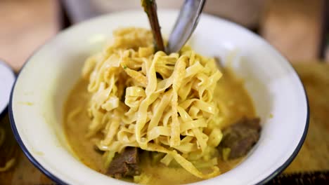 a bowl of northern thai beef curry noodles is stirred and lifted with chopsticks and a spoon under warm indoor lighting, highlighting rich textures and vibrant colors