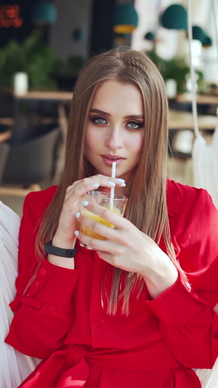 Portrait of sexy woman on a swing. Beautiful young lady drinking juice while sitting in a cafe and looking at camera. Vertical video