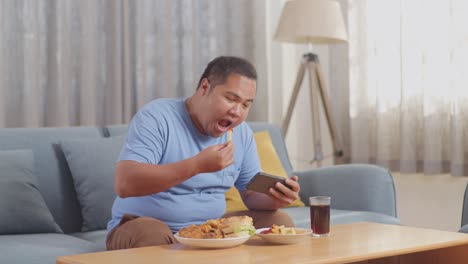 a fat asian man celebrating winning game on smartphone while eating fast food having french fries with ketchup on a sofa in the living room at home