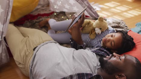 African-american-father-and-daughter-lying-in-tent-and-using-tablet-with-copy-space