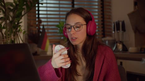 A-confident-and-serious-brunette-girl-in-red-wireless-headphones-and-glasses-looks-at-herself-in-the-mirror-and-studies-the-correct-pronunciation-in-practice-in-a-modern-apartment-in-the-kitchen-in-front-of-a-laptop-near-the-German-flag