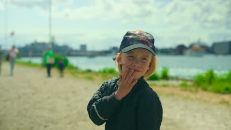 In-a-sunlit-urban-harbor,-a-7-year-old-boy-stands-with-a-straw-in-his-mouth-with-a-blurred-waterfront-behind-him