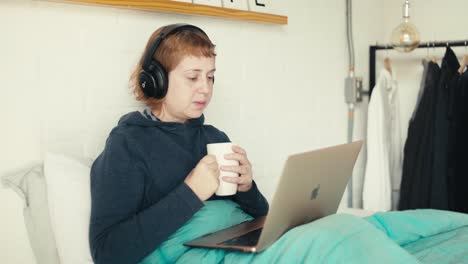 A-young-woman-sipping-warm-drink-while-viewing-laptop-from-bed