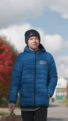 joven con chaqueta azul y gorra negra camina pensativo mirando a su alrededor en el área residencial, la luz del sol se refleja en su cara, con árboles de otoño y coche estacionado en el fondo