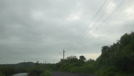 toma pov de un vehículo que viaja a lo largo de una carretera asfaltada con una línea eléctrica aérea que corre a lo largo de la carretera en un día nublado gris y sombrío, panjim, india