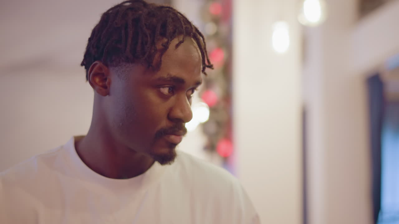 Man with braided hair in white shirt looks thoughtfully to side in festive room with holiday decorations, gazing calmly with lights and decorations in background