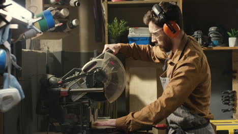 young caucasian male carpenter in goggles and headphones cutting wooden plank with a circular electric saw in carpentry workshop