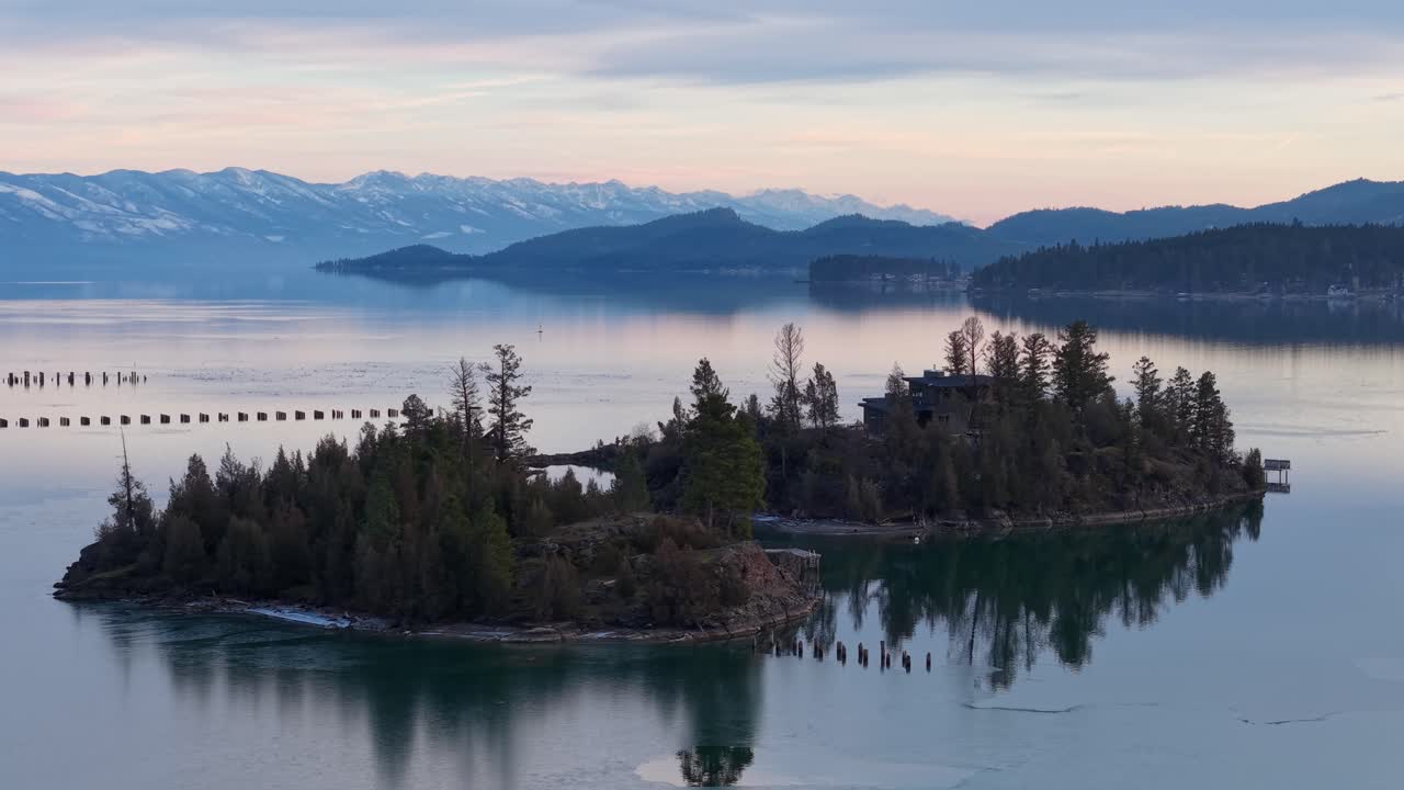 Veduta aerea pacifica di una piccola isola boscosa in un lago con montagne innevate in lontananza e acque calme.