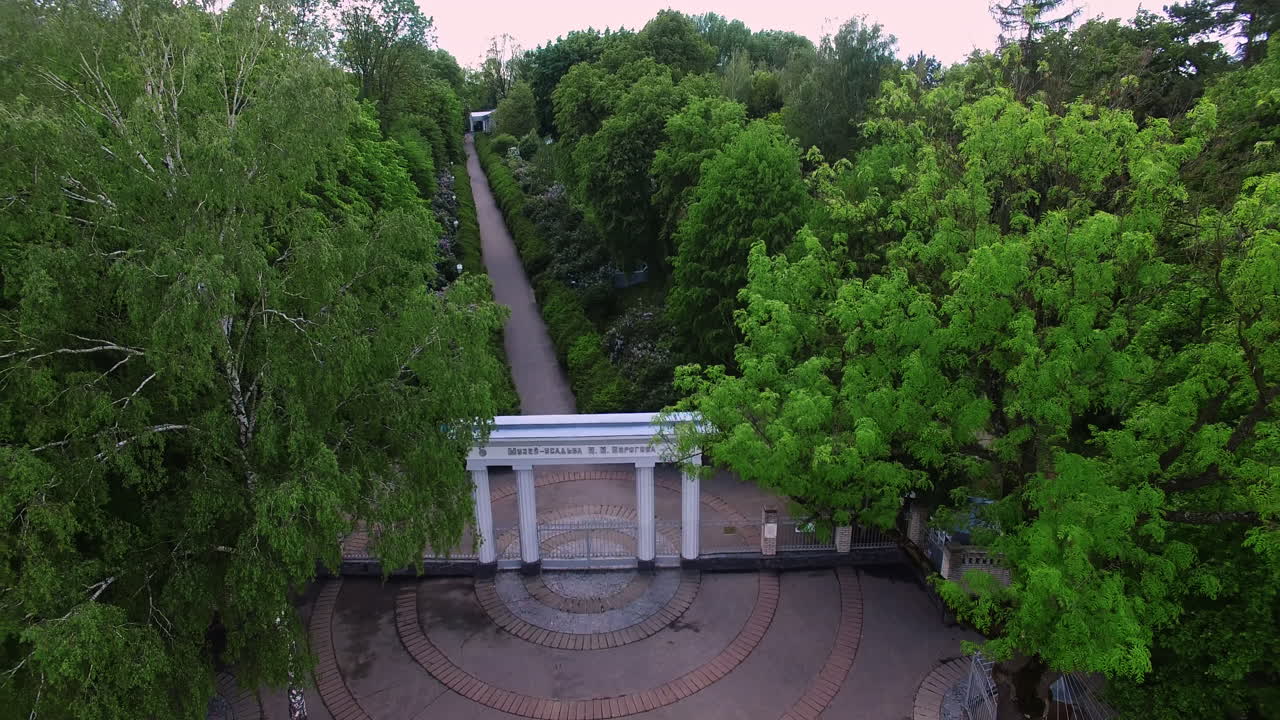Charming white entrance with columns to a green park. Drone rising over the beautiful territory of historical country seat with museum.