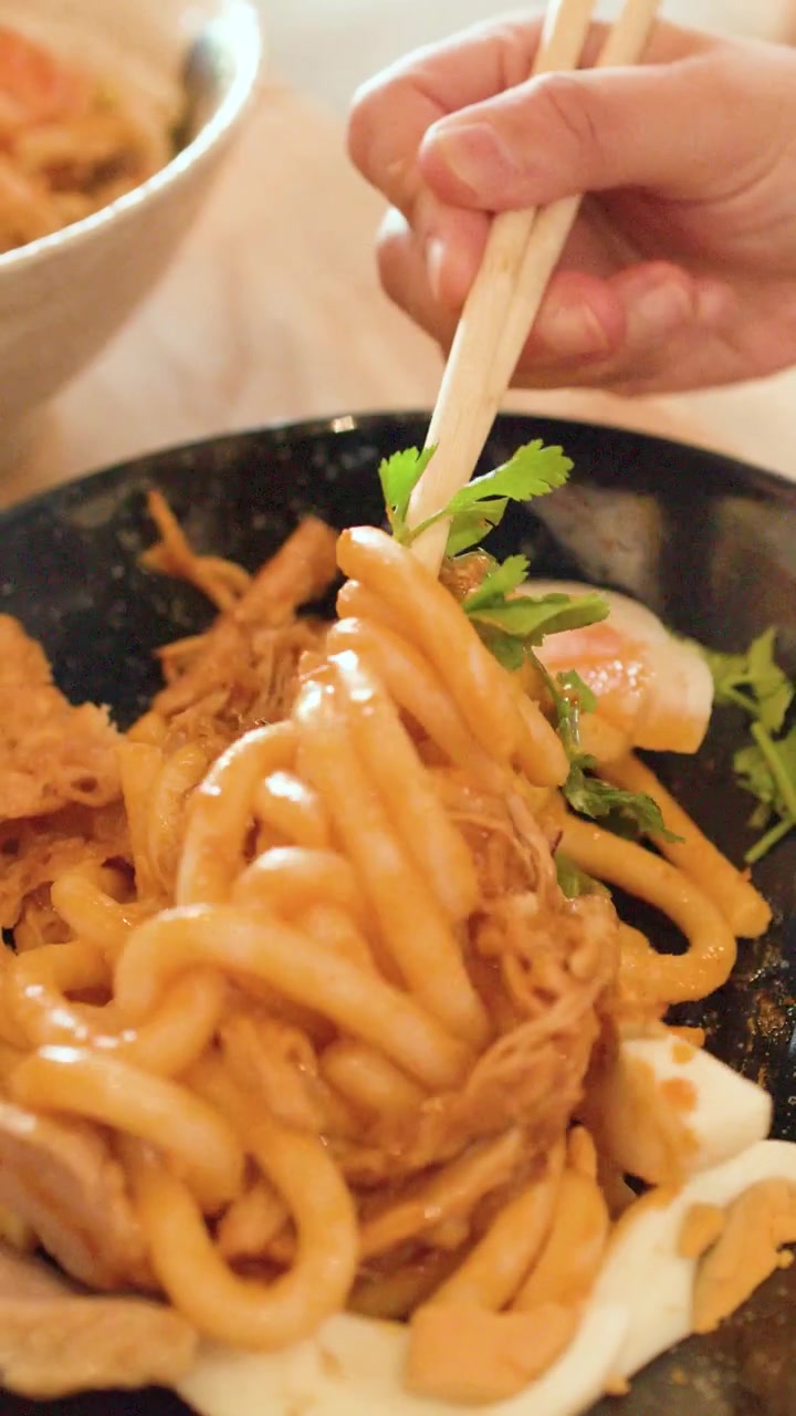 Primo piano di mani che mescolano spesse tagliatelle di riso con pollo e uovo usando bacchette di legno