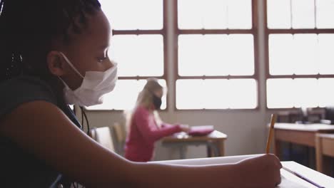 girl wearing face mask writing while sitting on her desk at school