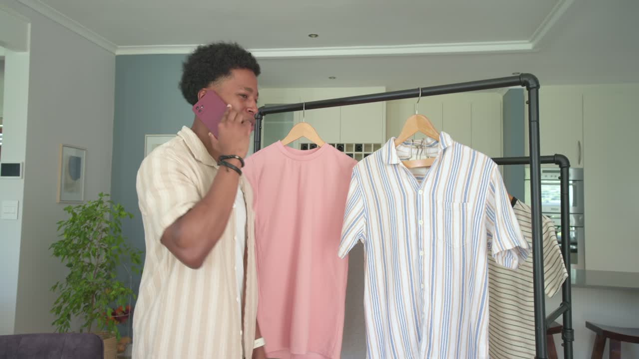 Uomo afroamericano adulto che telefona con un telefono magenta, toccando camicie rosa e blu e scegliendo un outfit a casa.