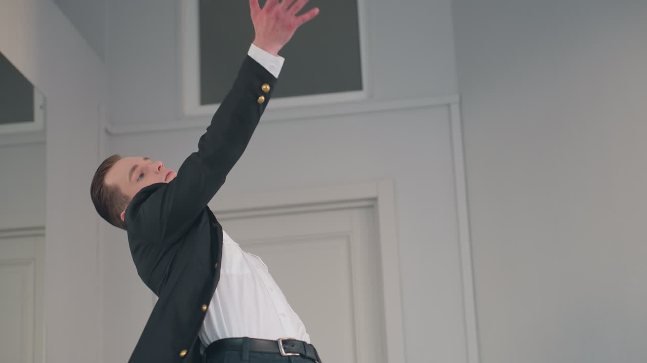 young man in black coat and white shirt walks forward then pivots into dramatic dance pose before mirror, expressive movement accentuated by minimalist studio backdrop and soft natural light
