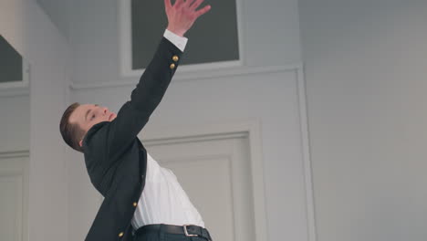 young man in black coat and white shirt walks forward then pivots into dramatic dance pose before mirror, expressive movement accentuated by minimalist studio backdrop and soft natural light