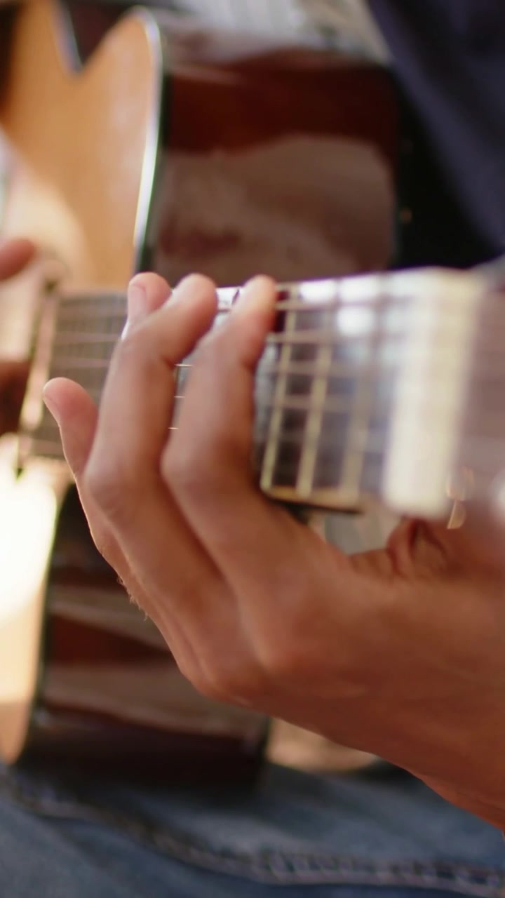 Vídeo vertical: Foco muda para um guitarrista formando acordes, tocando violão em casa, de jeans e jaqueta.