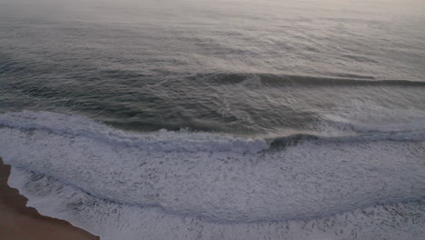 Espumosas-Olas-Rompiendo-En-La-Costa-Con-El-Antiguo-Paisaje-Del-Pueblo-En-La-Cima-De-Una-Colina-En-La-Playa-De-Nazare,-Portugal