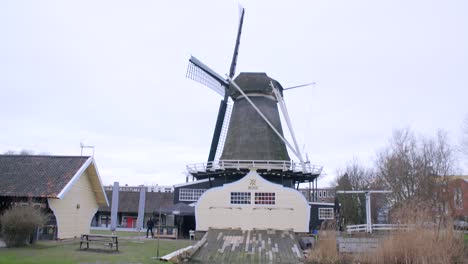 Peaceful-flowing-water-along-Utrecht-canal,-panning-up-to-a-large-wooden-windmill-and-jetty,-with-trees-and-rushes-blowing-in-the-wind