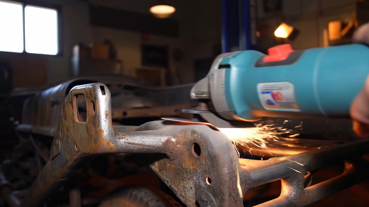 Close-up low-angle view shows handheld angle grinder sparking against corroded vehicle chassis bracket inside dim garage workshop, highlighting rough metal texture, repair task, and industrial setting