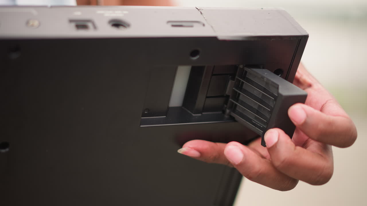 Nærbillede af hånd, der fjerner batteripakke fra rum, fingre trækker lille strømmodul ud fra bageste åbning på elektronisk enhed, synlige stik og plastlås, præcis bevægelse, udendørs.